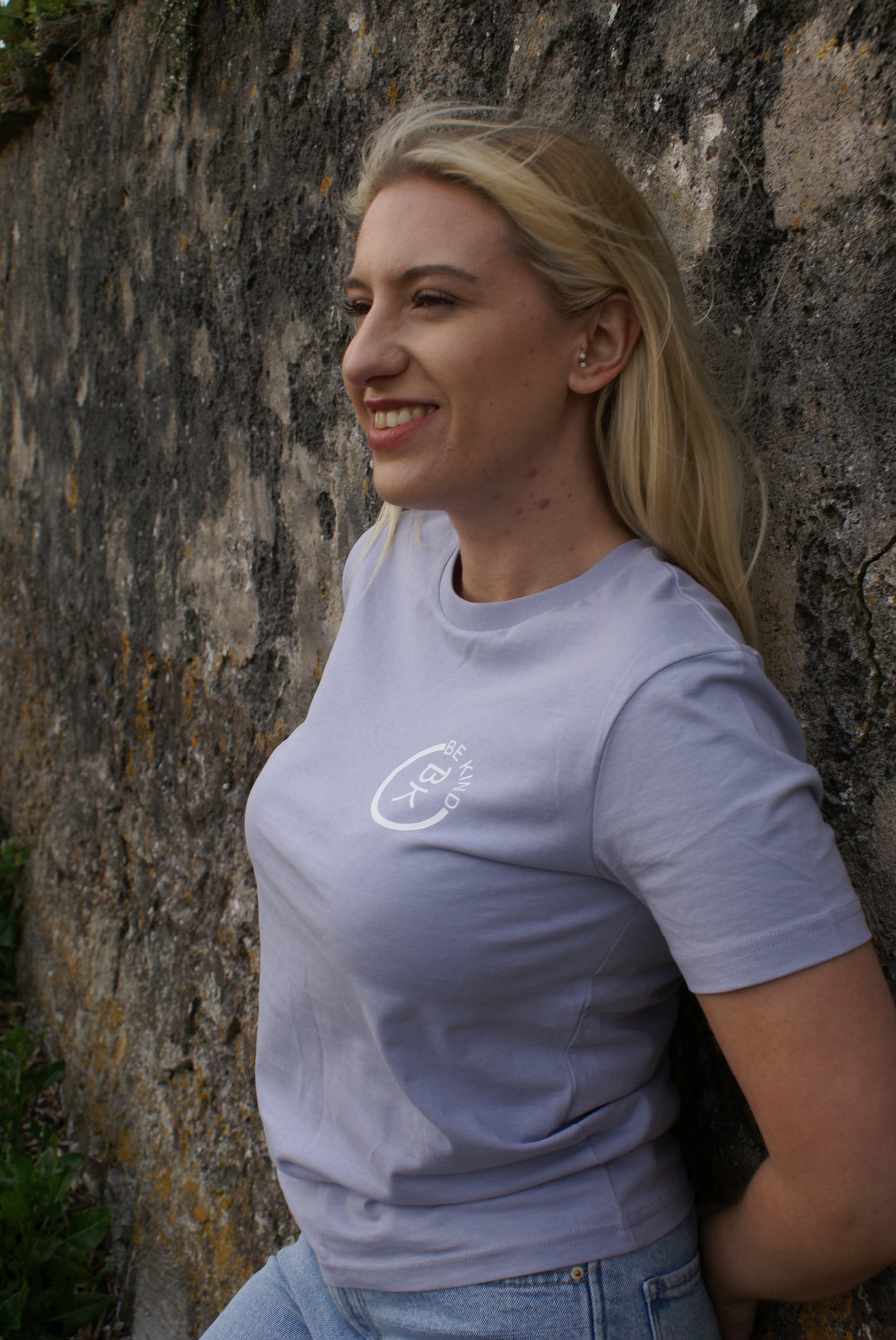 A woman leans on a wall, she's wearing a Lavender Organic Cotton T-Shirt from the Be Kind Apparel Original Collection