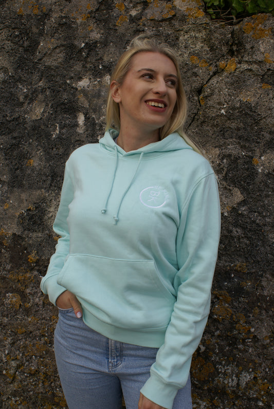A woman stands by a rock wall, she's wearing a Mint Green Organic Cotton Hoodie from the Be Kind Apparel Original Collection