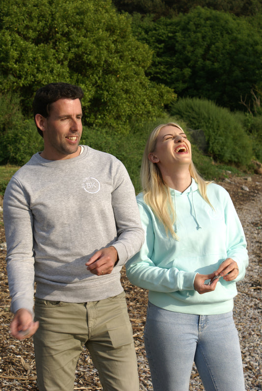 A man stands next to a woman on a beach, he's wearing a Grey Organic Cotton sweatshirt from Be Kind Apparel's Freestyle Range