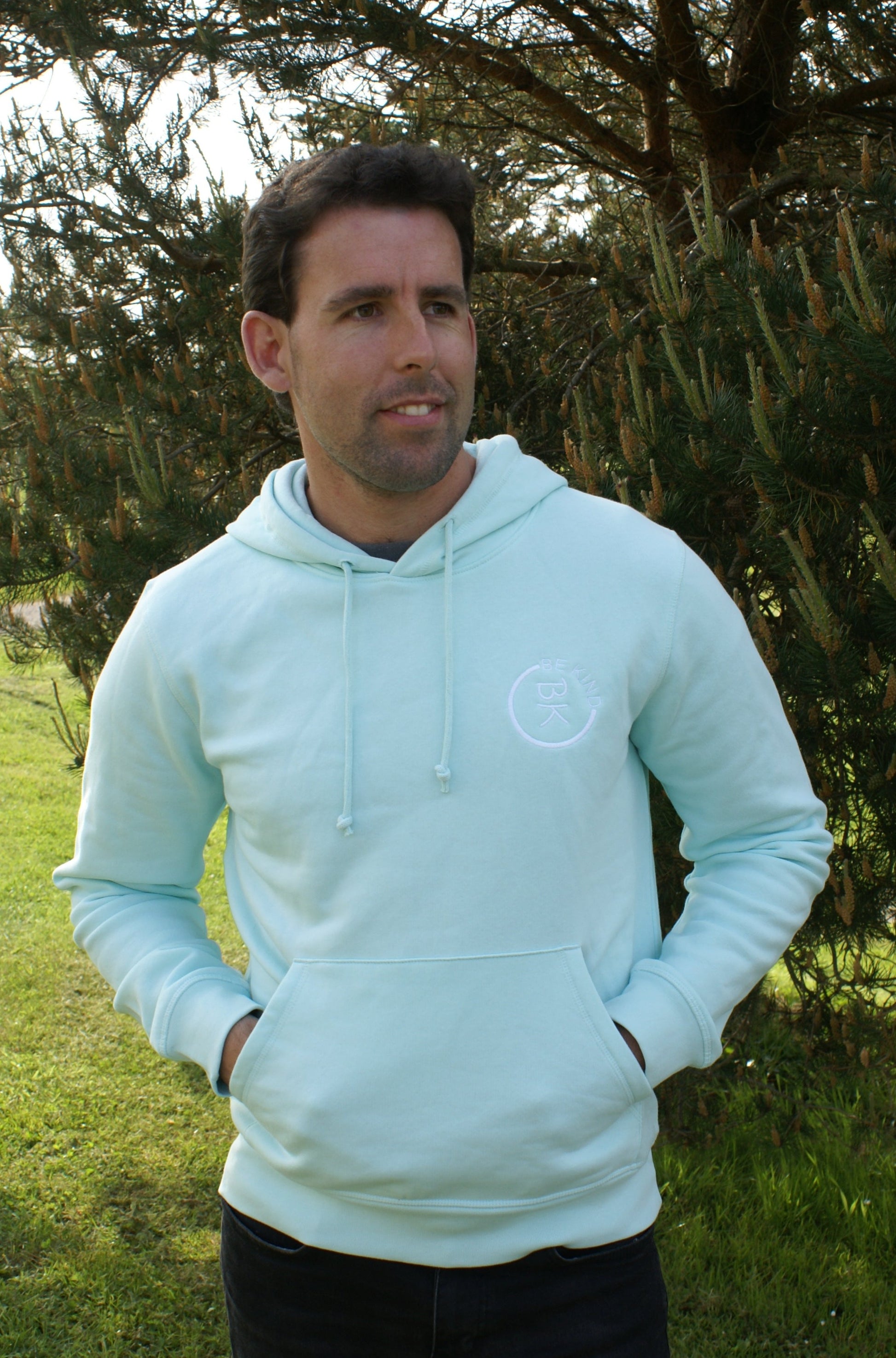 A man stands by a tree, he's wearing a Mint Green Organic Cotton Hoodie from the Be Kind Apparel Original Collection