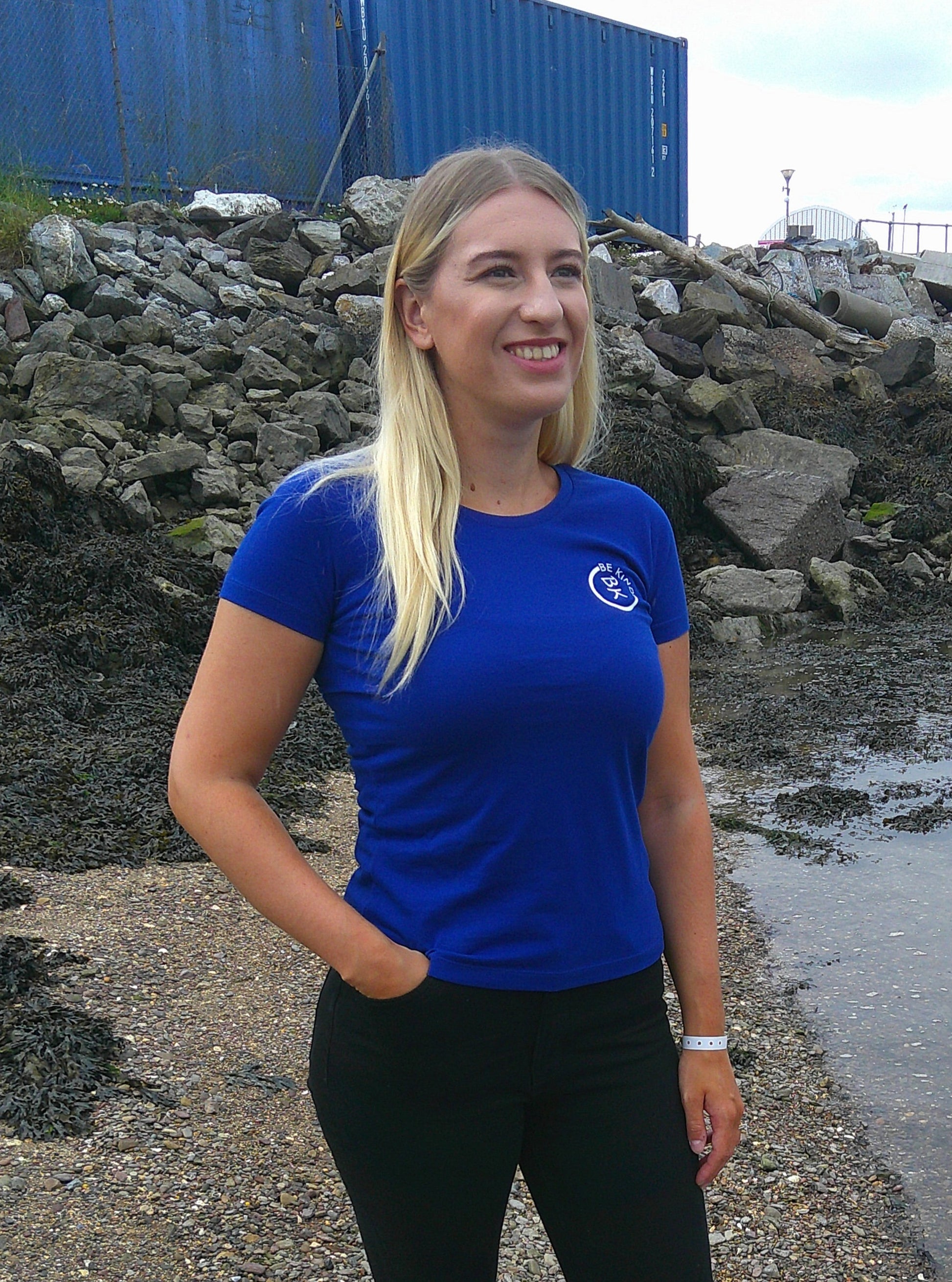 A woman stands on a beach, she's wearing a Cobalt Blue Organic Cotton Connector T-Shirt from the Be Kind Apparel Connector Range