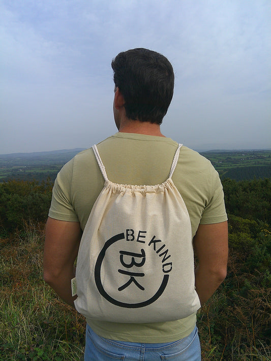 A man looks away to the hills, he's got an 100% Organic Canvas drawstring bag on his back with the official Be Kind Apparel logo in black