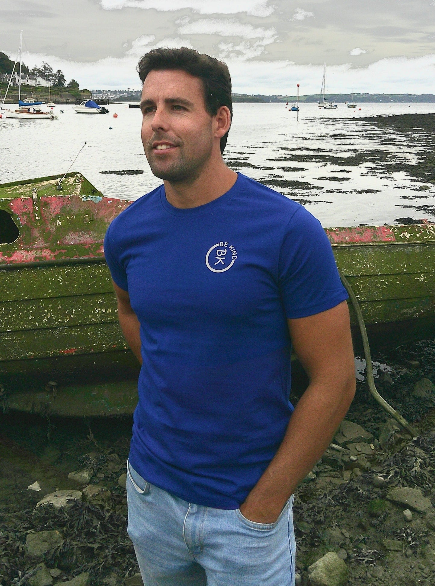 A man stands by a boat, he's wearing a Cobalt Blue Organic Cotton Connector T-Shirt from the Be Kind Apparel Connector Range
