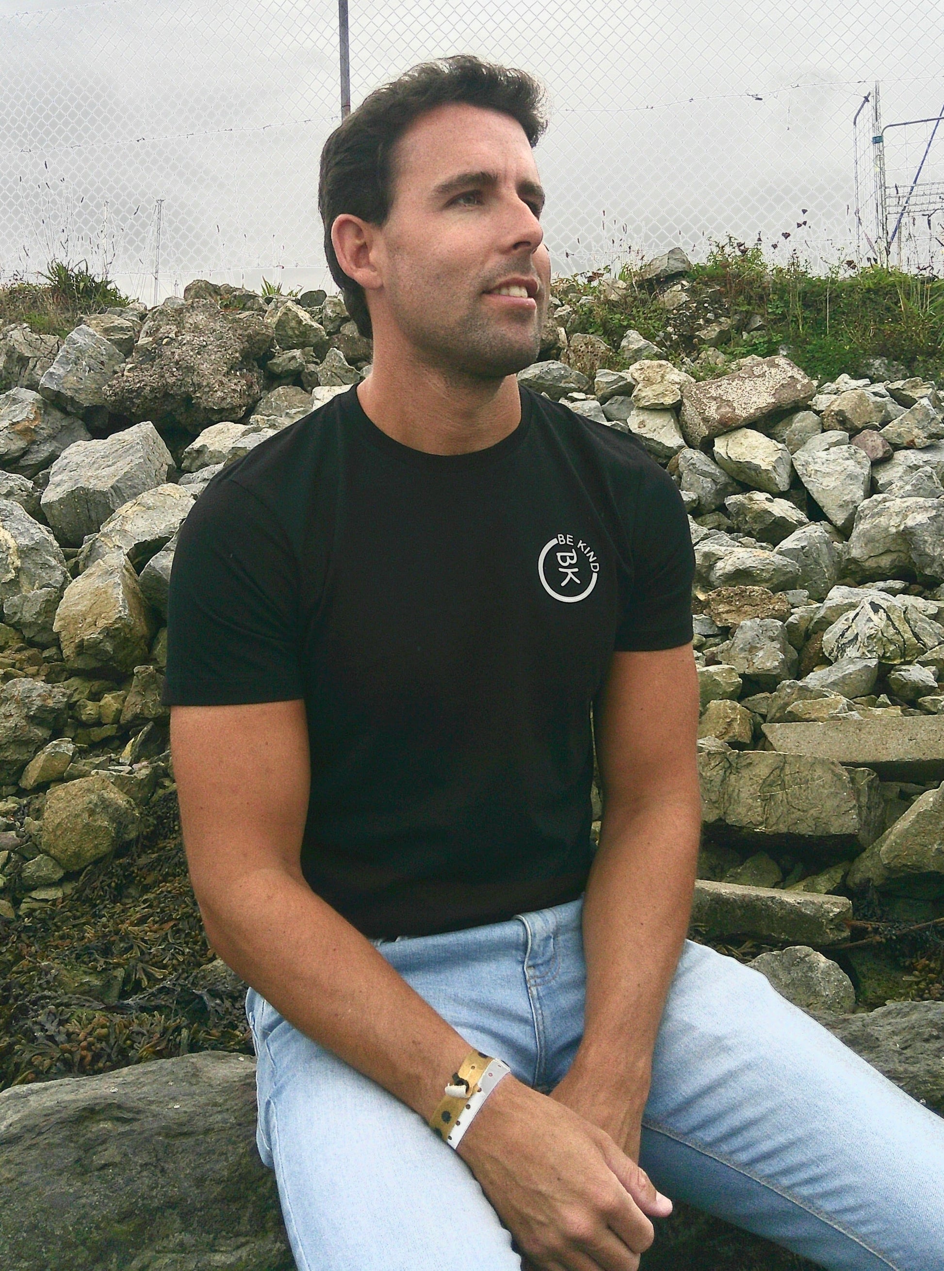 A man sits, he's wearing a Black Organic Cotton Connector T-Shirt from the Be Kind Apparel Connector Range