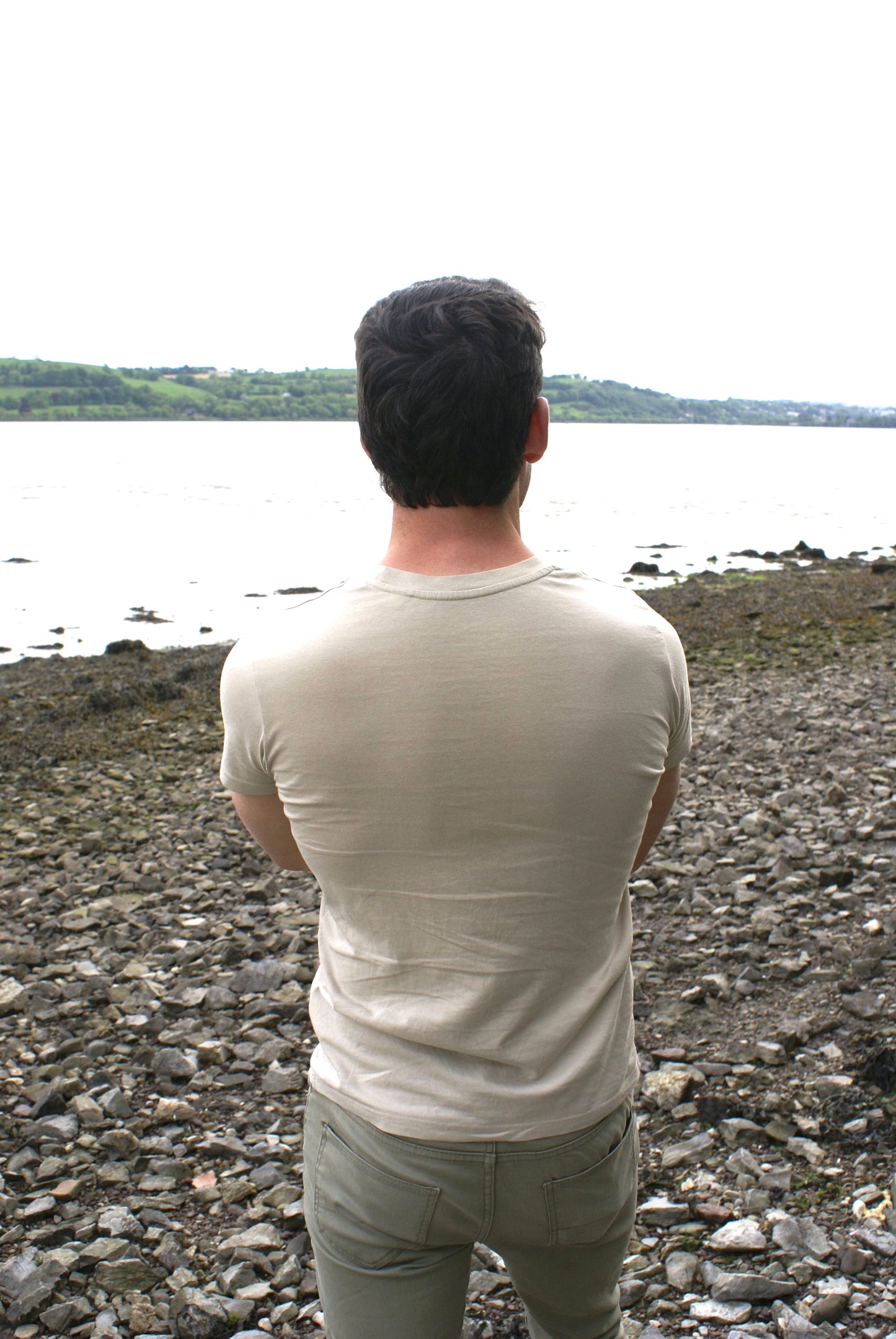 A man looks out at a river, he's wearing a Sandstone Beige Organic Cotton T-Shirt from the Be Kind Apparel Original Collection