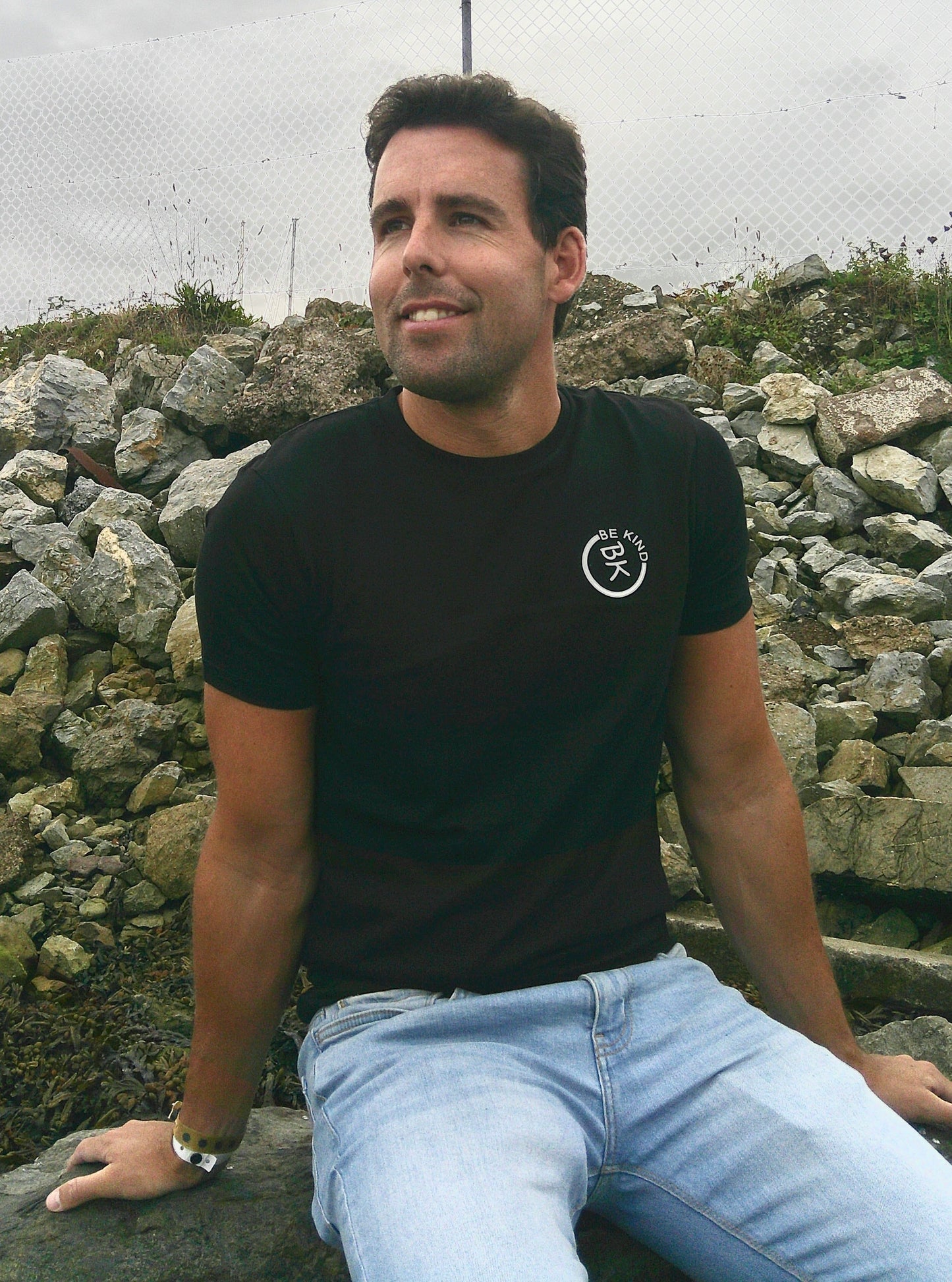 A man sits by the shore, he's wearing a Black Organic Cotton Connector T-Shirt from the Be Kind Apparel Connector Range