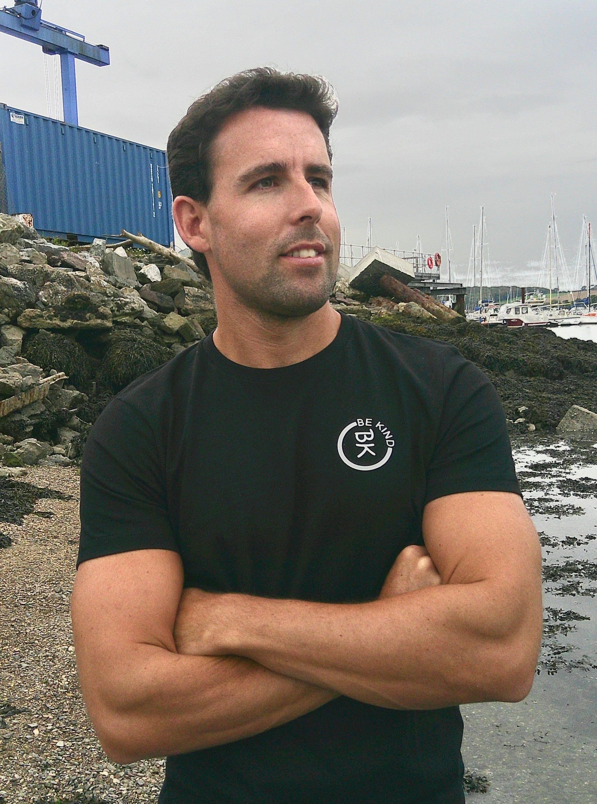 A man stands with his arms crossed, he's wearing a Black Organic Cotton Connector T-Shirt from the Be Kind Apparel Connector Range