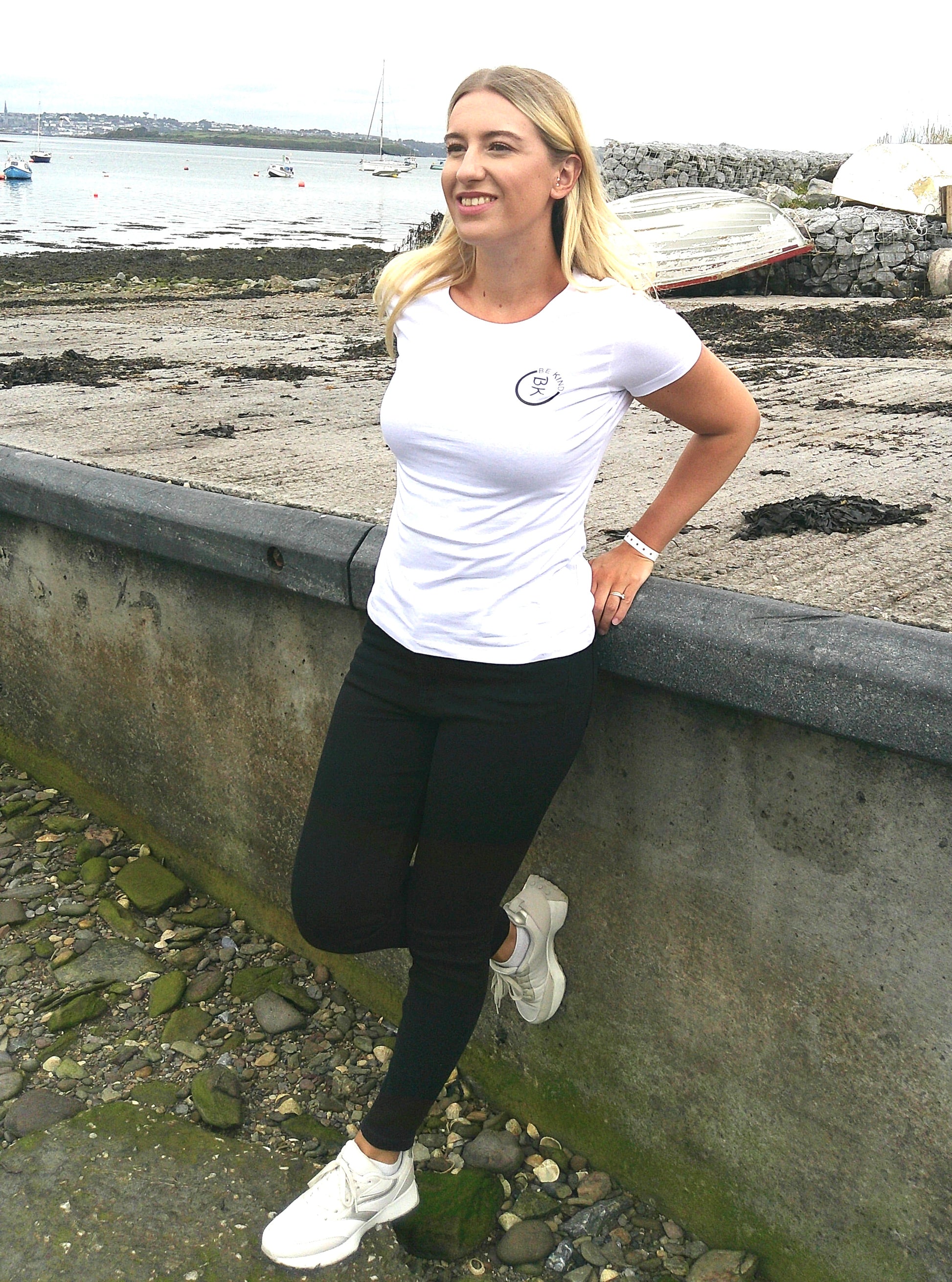 A woman leans against a wall, she's wearing a White Organic Cotton Connector T-Shirt from the Be Kind Apparel Connector Range