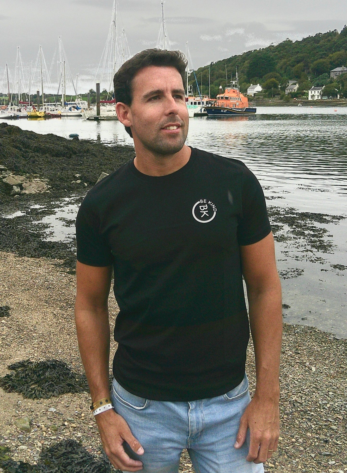 A man stands on a beach, he's wearing a Black Organic Cotton Connector T-Shirt from the Be Kind Apparel Connector Range