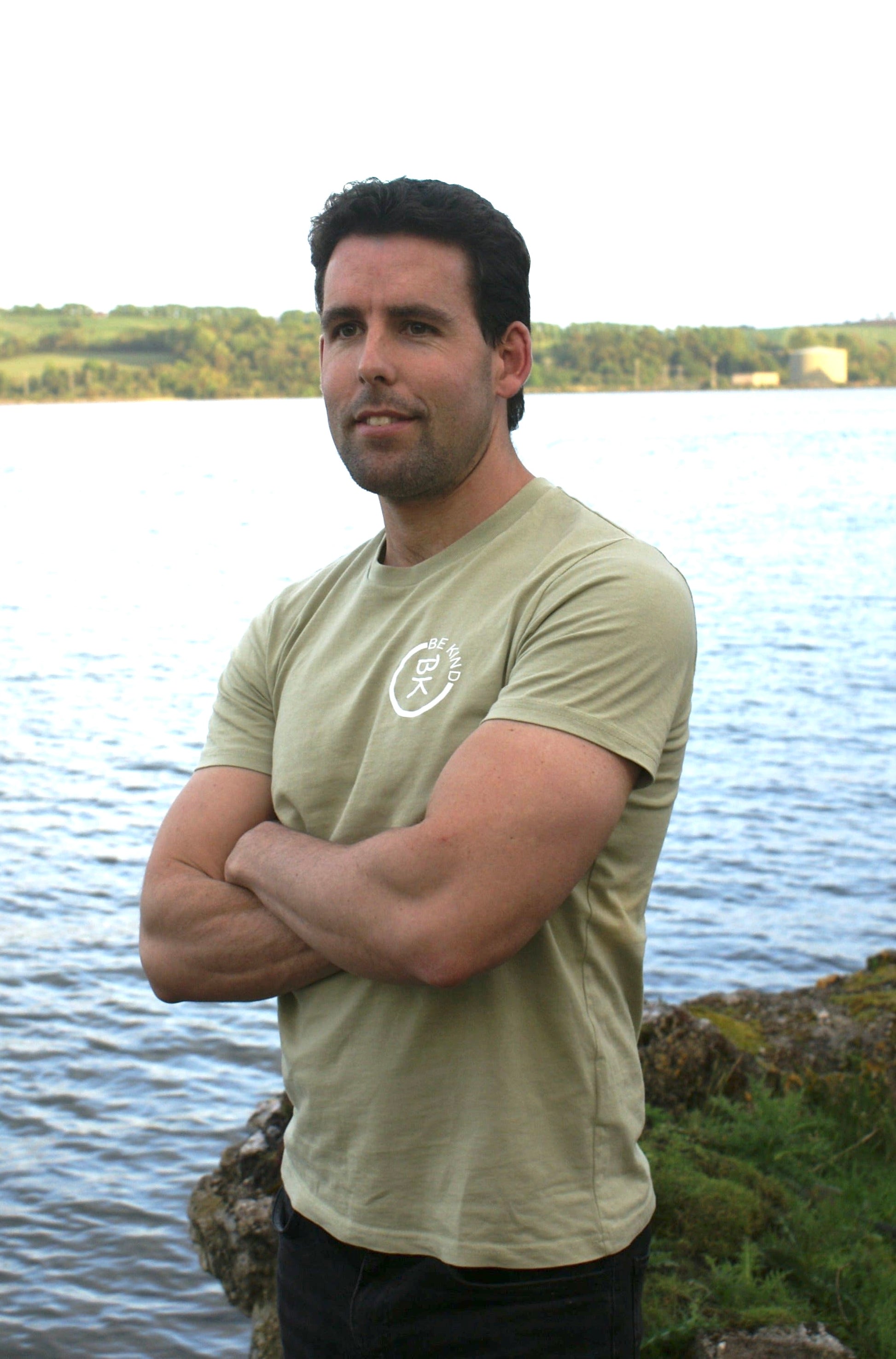 A man stands by the shore with folded arms, he's wearing an Olive Green Organic Cotton T-Shirt from the Be Kind Apparel Original Collection