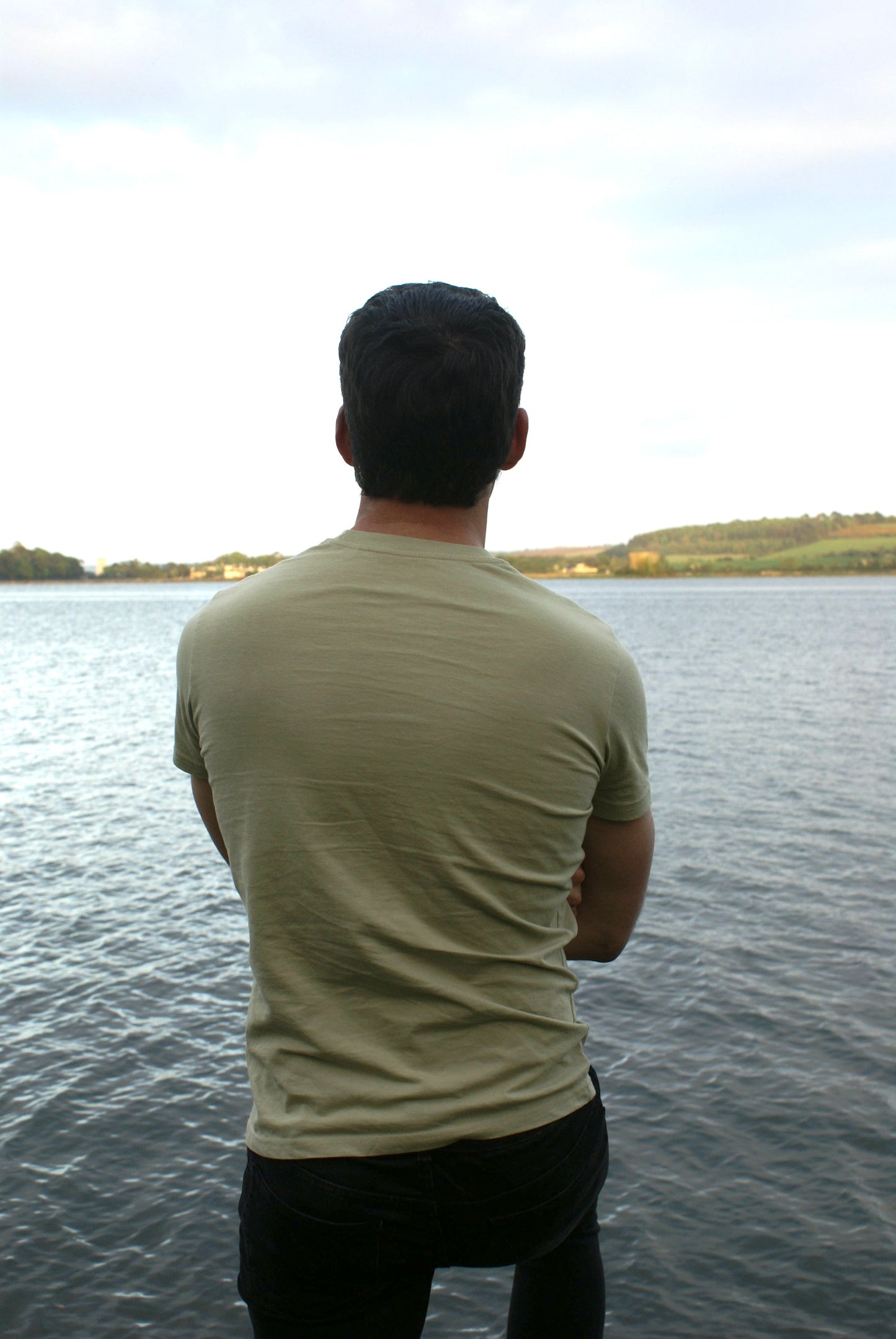 A man looks out across the water, he's wearing an Olive Green Organic Cotton T-Shirt from the Be Kind Apparel Original Collection