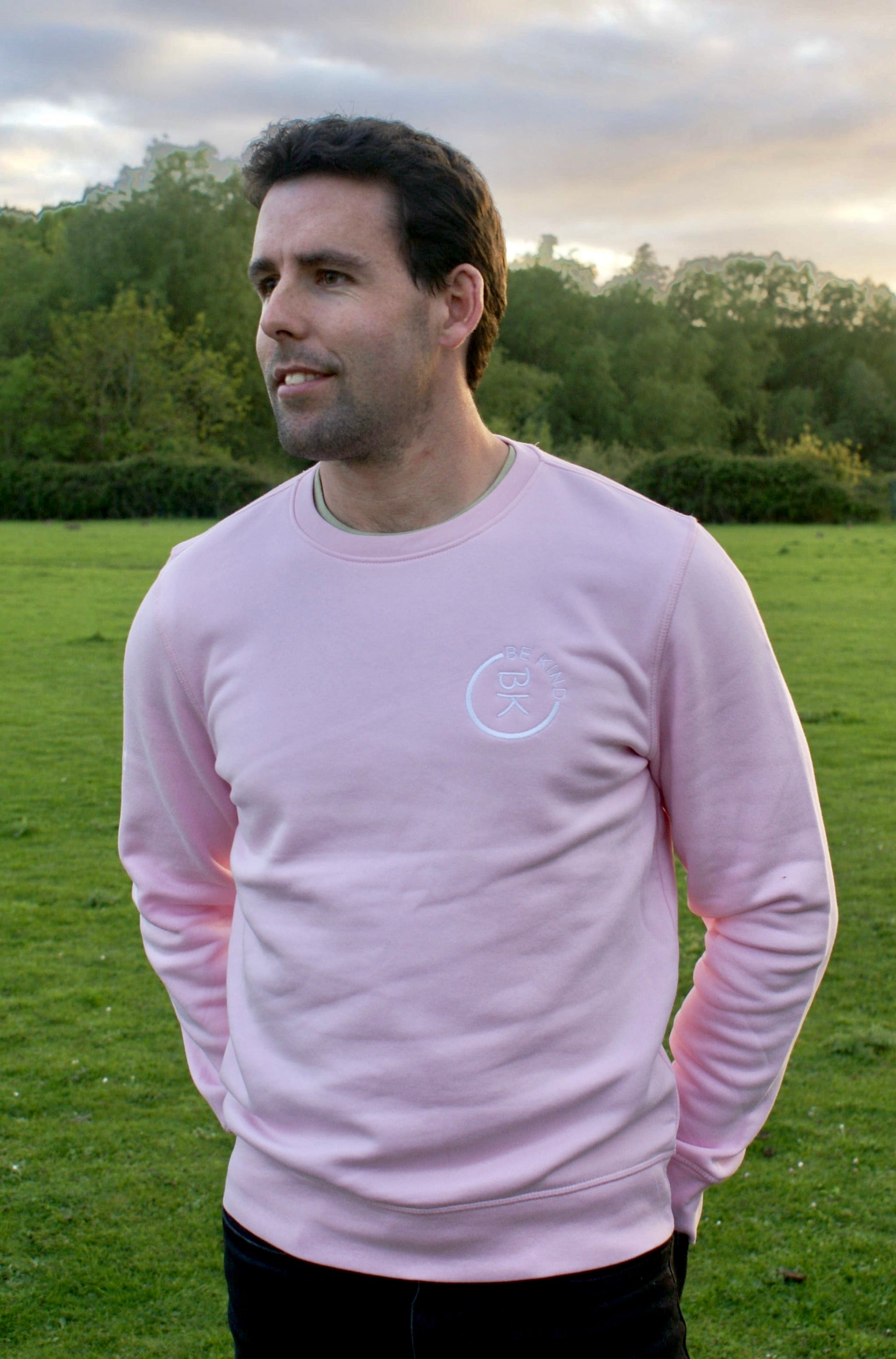 A man stands in a green field, he's wearing a Bubblegum Pink Organic Cotton Sweatshirt from the Be Kind Apparel Original Collection